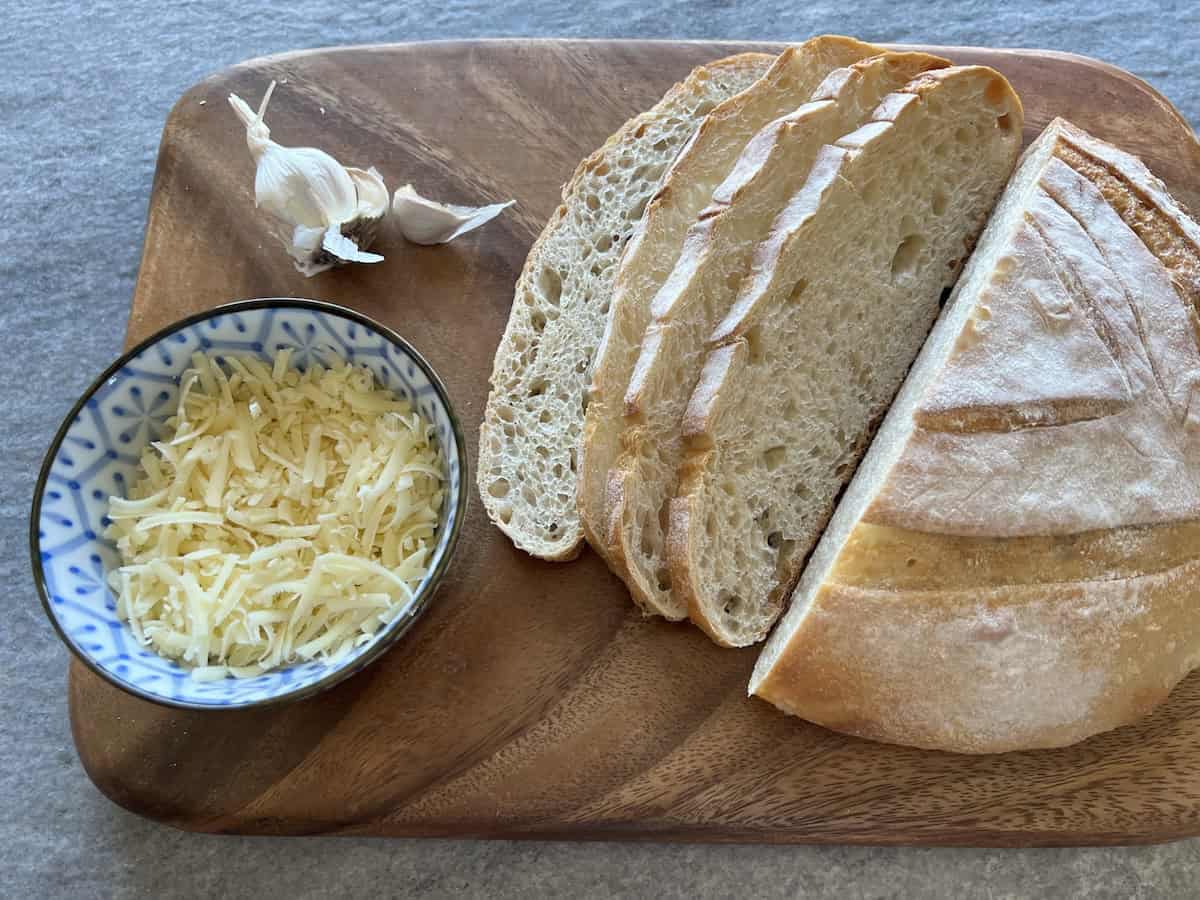 a wooden board with sliced sourdough bread, whole garlic cloves, and a bowl of shredded gruyere cheese on it.