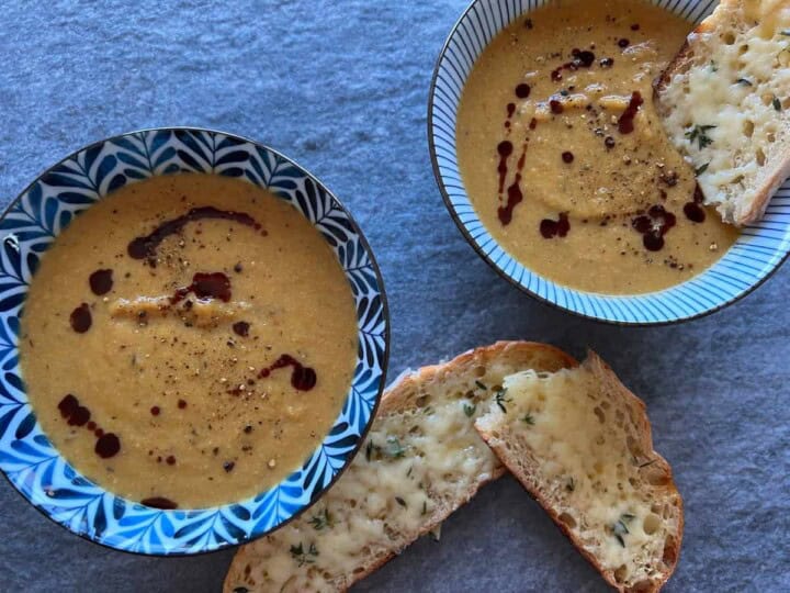 two bowls of creamy, orange colored pumpkin soup set on a stone surface. the soup is garnished with dots of dark brown pumpkin seed oil, black pepper, and cheesy thyme toast.