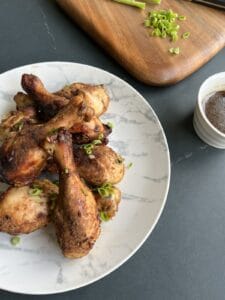 golden brown crockpot chicken legs on a platter, next to chopped scallions on a cutting board and a small bowl of honey-garlic soy sauce