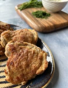 crispy pickle brine chicken thighs on a serving platter, with fresh chopped dill and a bowl of sauce on a wooden board in the background.