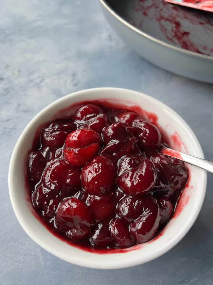 dark red cherry compote in a white bowl. the cherry compote has a glossy texture and the pan it was cooked in sits in the background