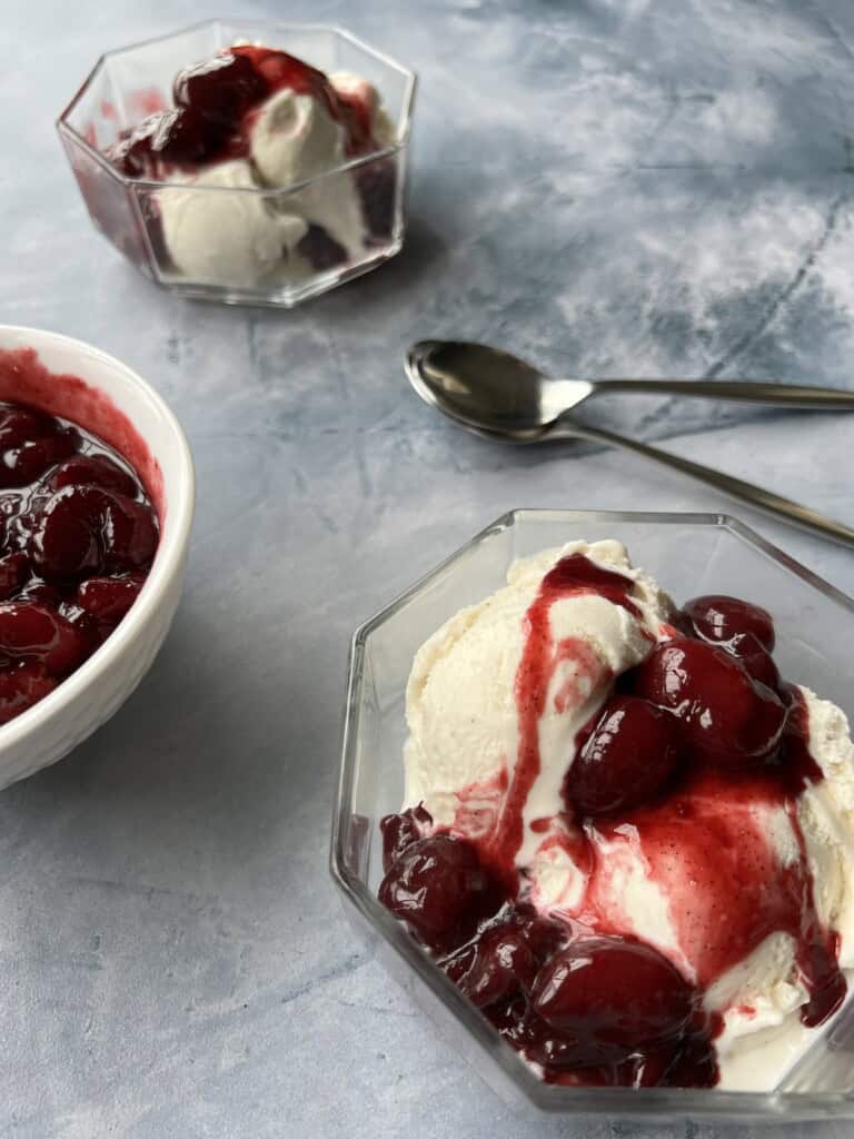 2 geometric glass bowls of vanilla ice cream topped with cherry compote. the cherry compote features whole cherries suspended in a thick red juice