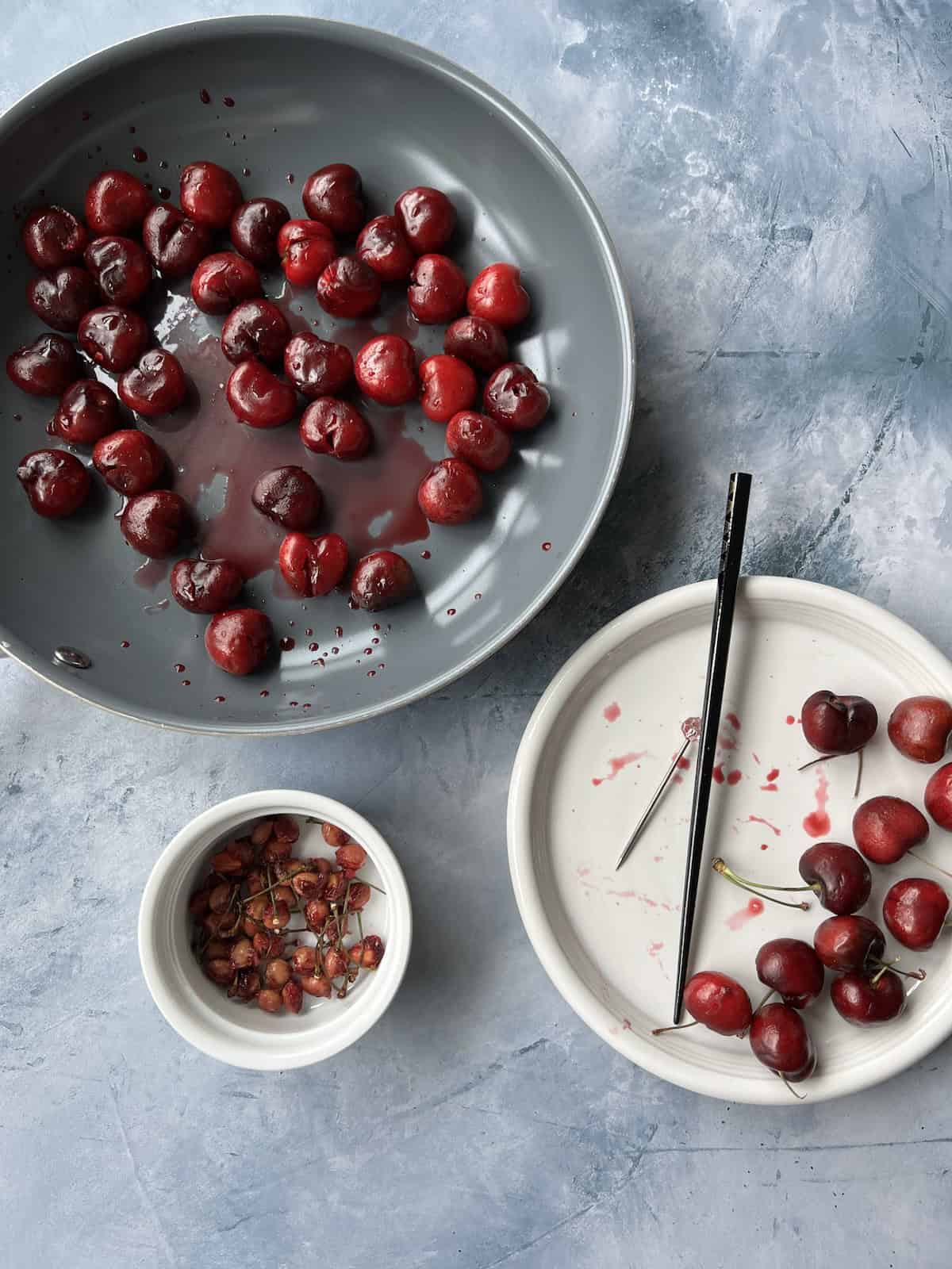 a pan with pitted cherries is next to a plate with a few whole cherries. there is also a small bowl containing cherry pits and stems, as well as the tools used  to pit the cherries. 