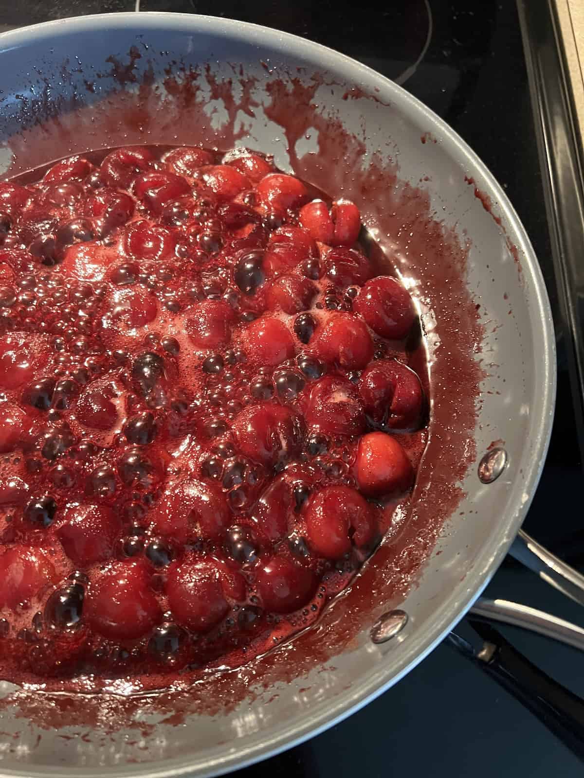 the cherry mixture being cooked in the skillet. the cherry juices are producing foamy bubbles as the cook and steam is rising from the pan. 