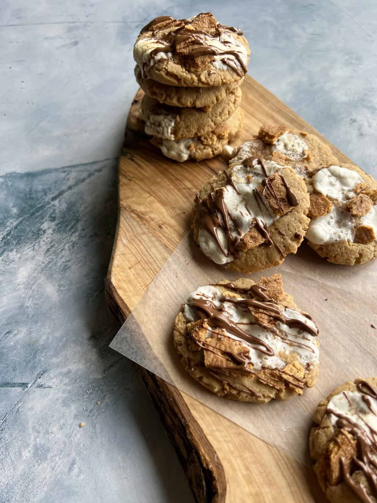 peanut butter s'mores cookies on a wooden serving board. one cookie is front and center to show off its chocolate drizzle and there is a tall stack of cookies in the background.