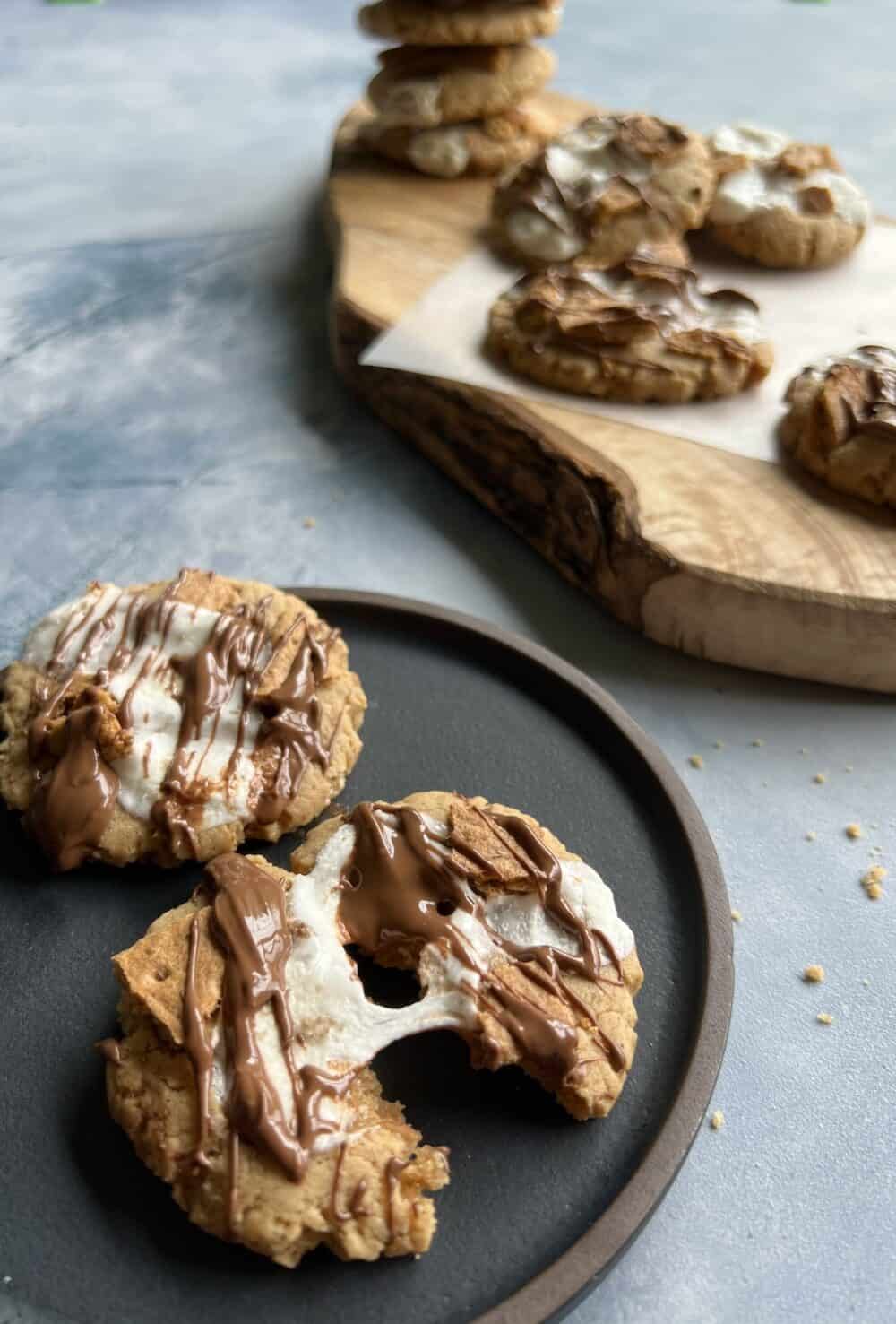 two peanut butter smores cookies on a dark matte plate. one of the cookies is partially split, showing off the gooey texture of the marshmallow as it pulls apart.