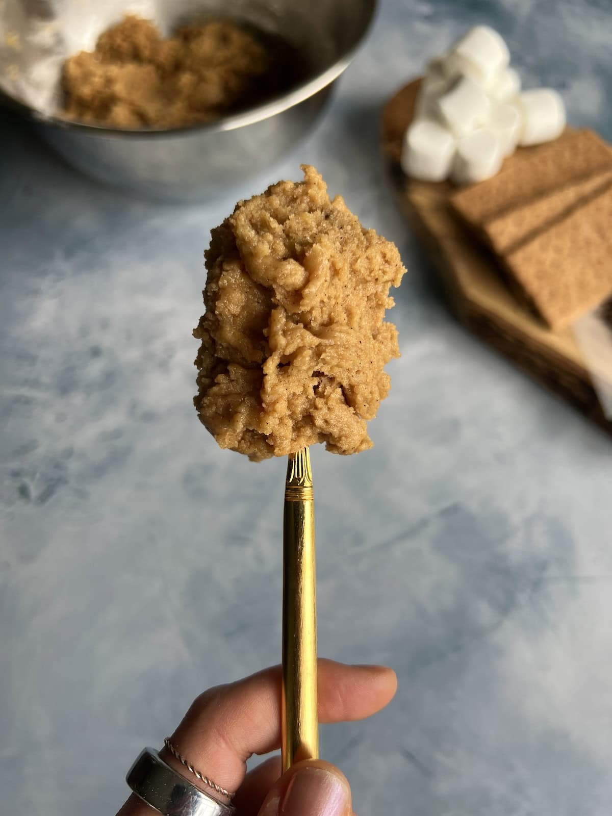 a spoon bearing a glob of peanut butter s'more cookie dough is held close to the camera, showing off its thick and sticky texture. in the background are a bowl containing more cookie dough and a rustic wooden board with s'more toppings. 