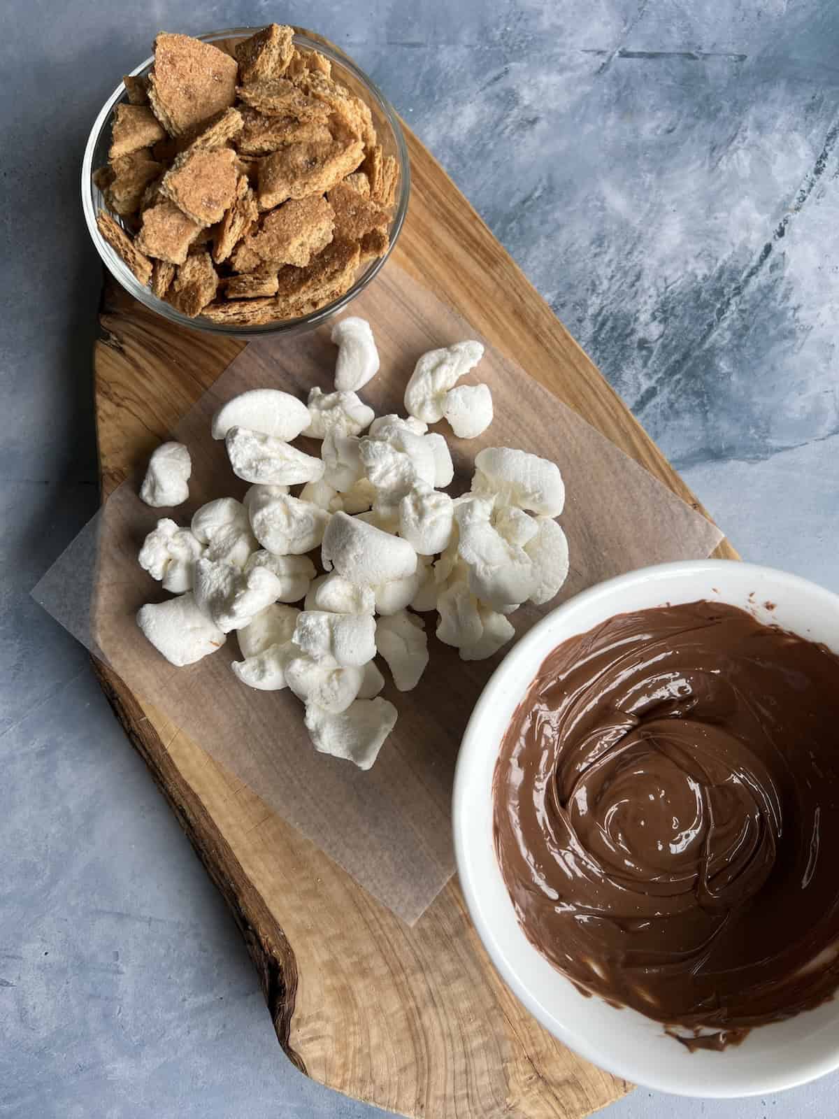 crumbled graham crackers, torn marshmallow bits, and a bowl of melted chocolate arranged on a rustic wooden board.  
