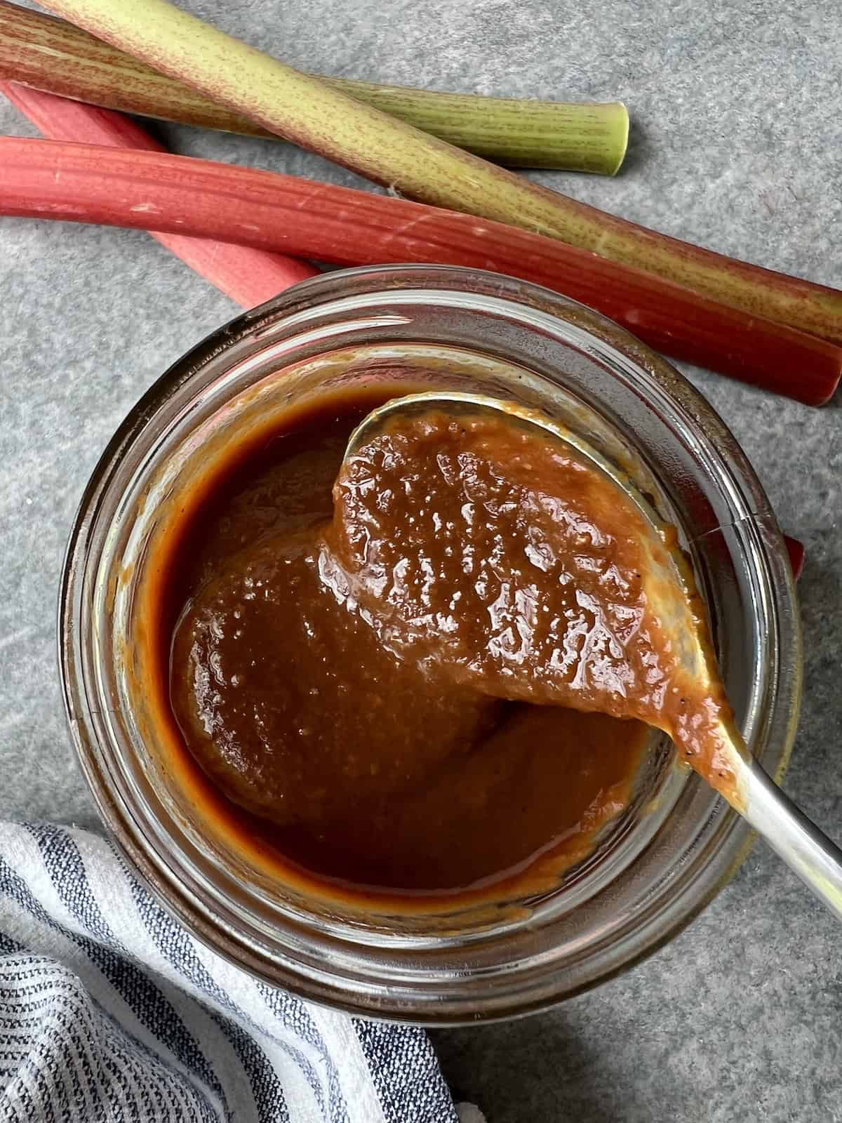 rhubarb bbq sauce in a mason jar. a spoon is being dipped into the sauce, show its thick and shiny texture.