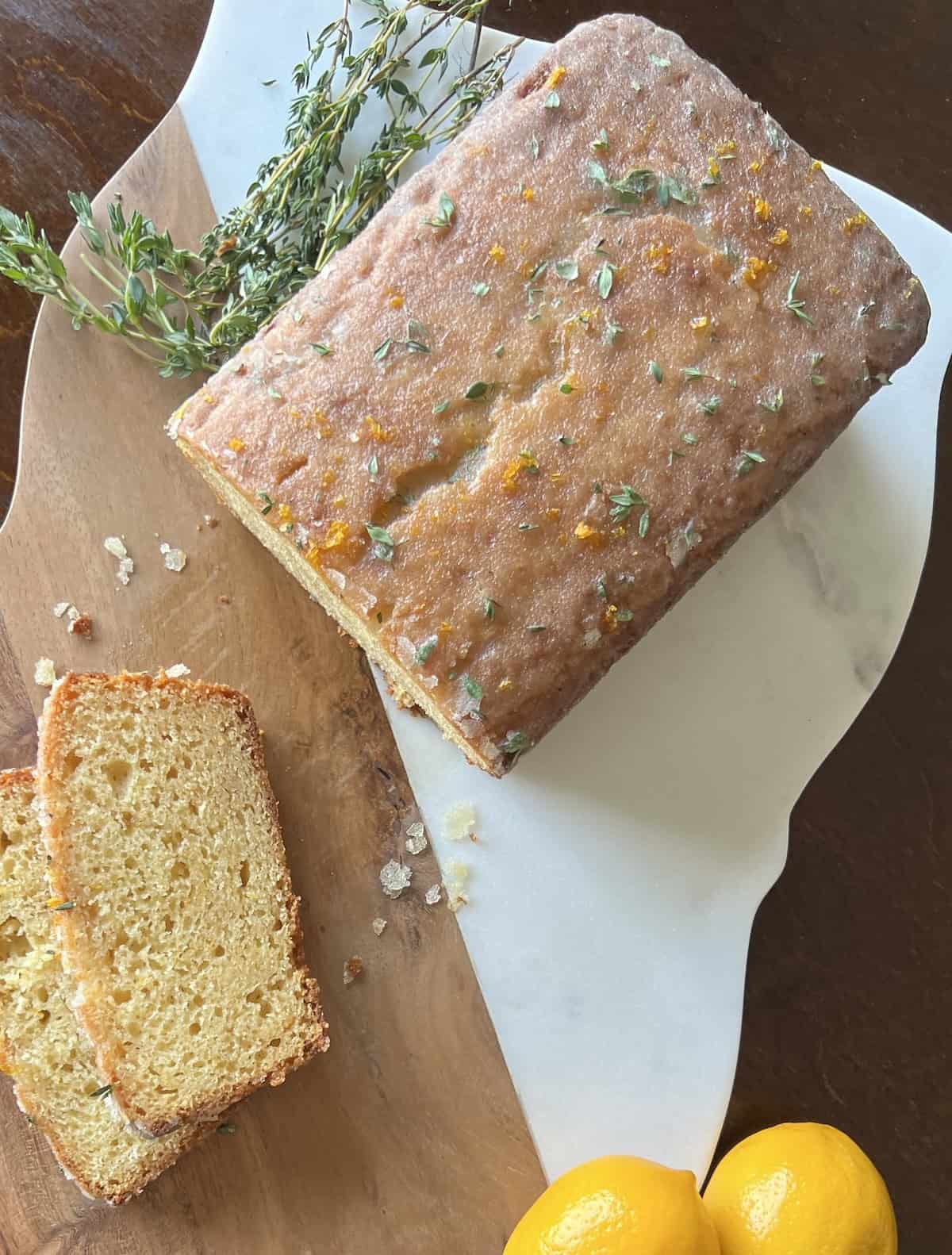 whole meyer lemon cake sits on a cutting board. 2 slices have been cut off and fresh thyme sprigs and whole lemons are nearby.