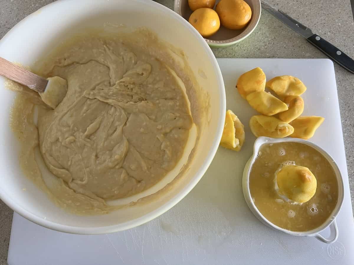 a white glass bowl to the left containing the egg mixture and the flour mixture, blended together. to the right, a citrus juicer containing the juice of 4 meyer lemons, with the already squeezed fruit rinds in a pile next to it.