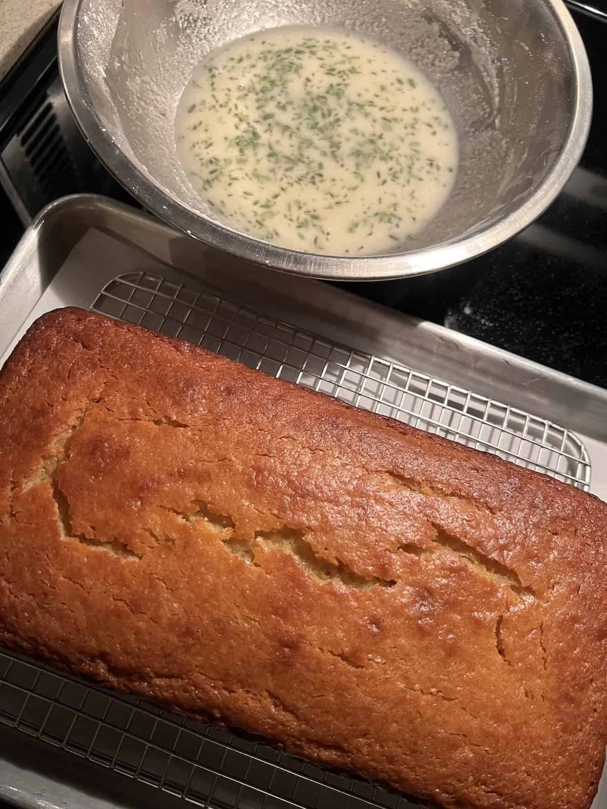 the baked meyer lemon loaf cake sitting on a metal wire rack that is positioned on top of a baking sheet. a stainless steel bowl containing a meyer lemon juice and thyme glaze is sitting next to the cake.