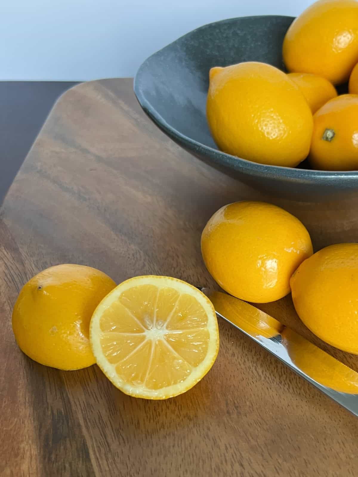 a sliced Meyer lemon sits on a cutting board facing the camera, showing off the juicy and brightly colored interior. a bowl of Meyer lemons and a chef's knife sit next to it.