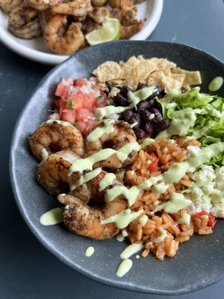 a stone bowl filled with charred shrimp, Spanish rice, black beans, pico de gallo, and various other toppings. a plate with more charred shrimp and lime wedges sits in the bakground.