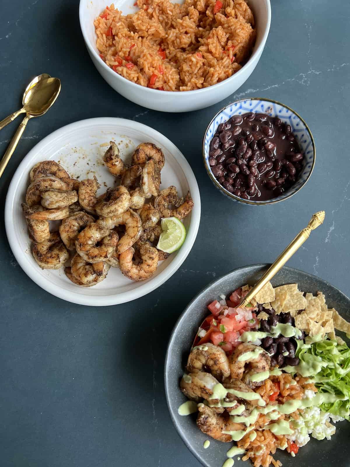 a finished shrimp burrito bowl sits on a countertop alongside a plate of seared shirmp, a small bowl of black beans, and a large bowl of orange-colored spanish rice.
