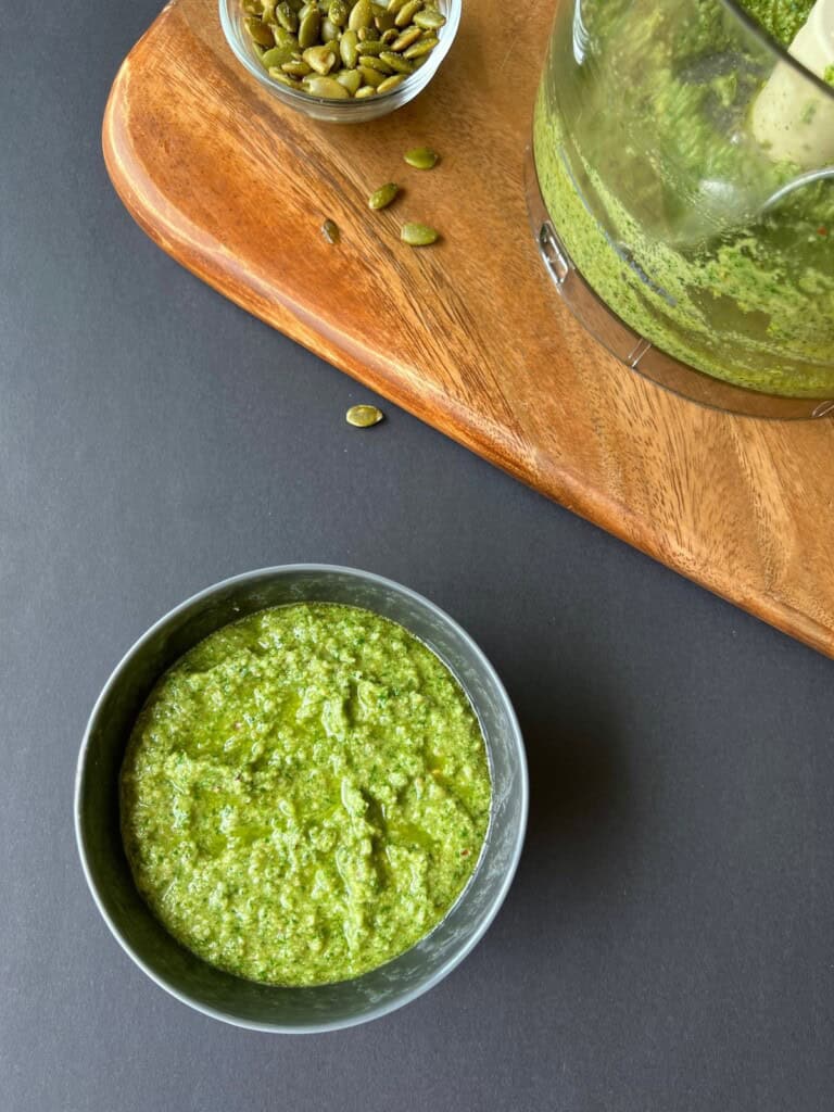 pumpkin seed pesto is transferred from the bowl of a food processor to a small bowl for serving