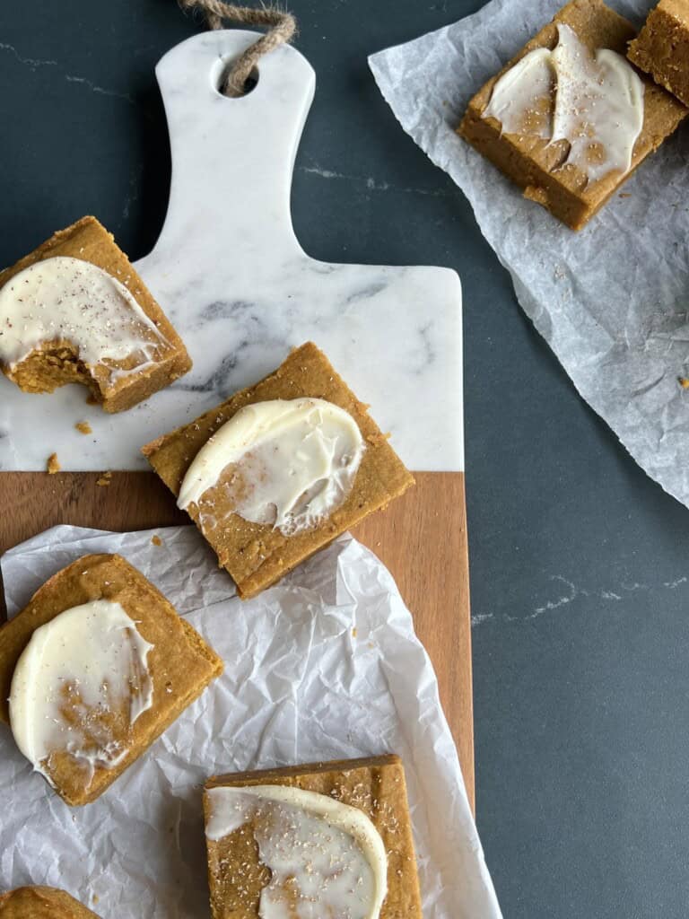 frosted pumpkin spice blondies are arranged on a two tone cutting board