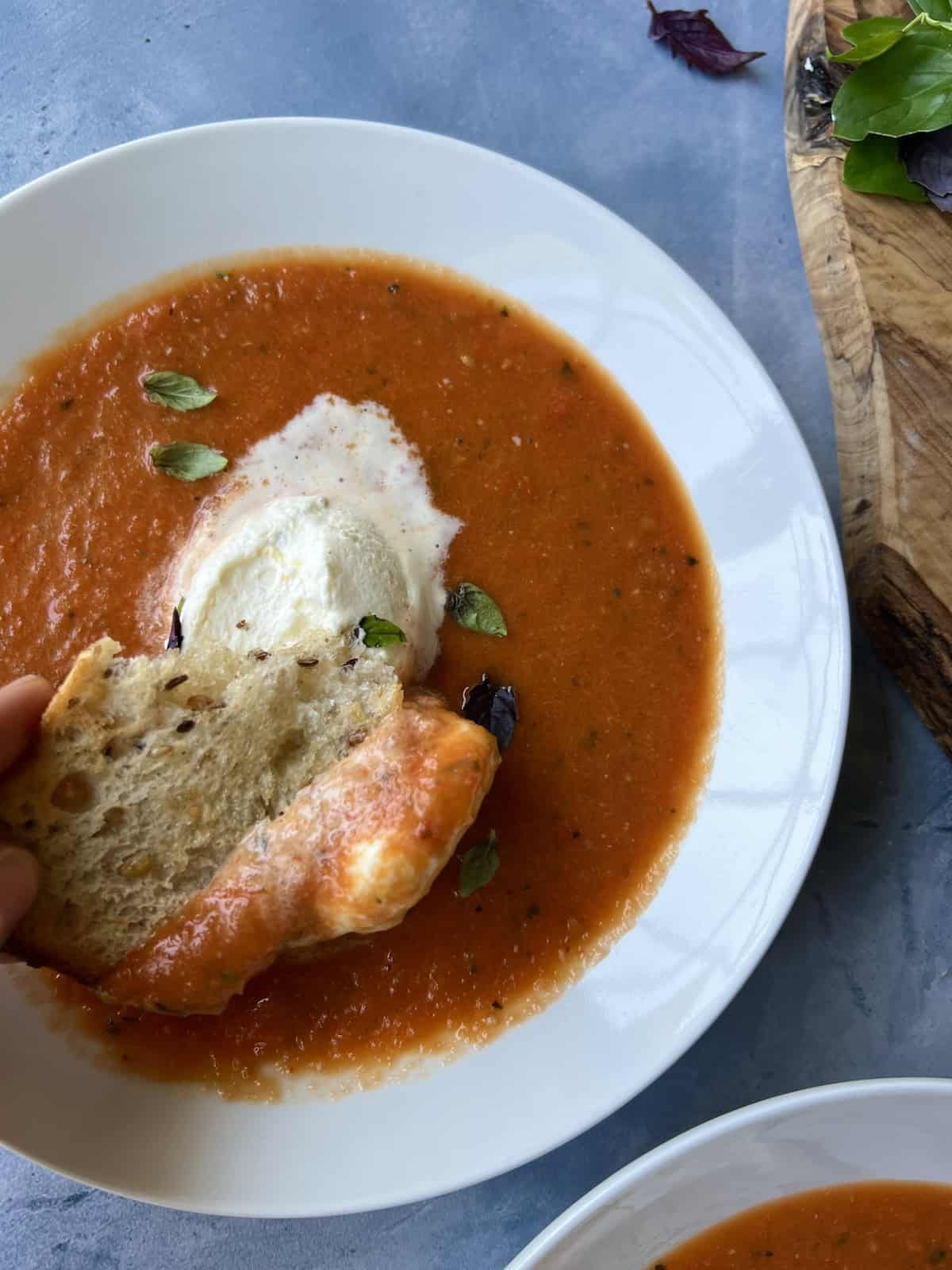 a piece of crusty bread is dipped into the savory whipped cream garnish atop a bowl of roasted garlic and tomato soup