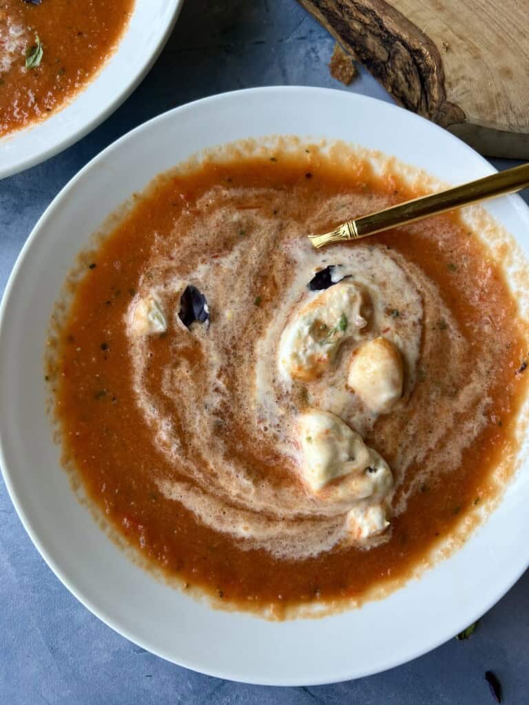 savory whipped cream is stirred into a bowl of roasted garlic and tomato soup, creating swirls of deep red and white