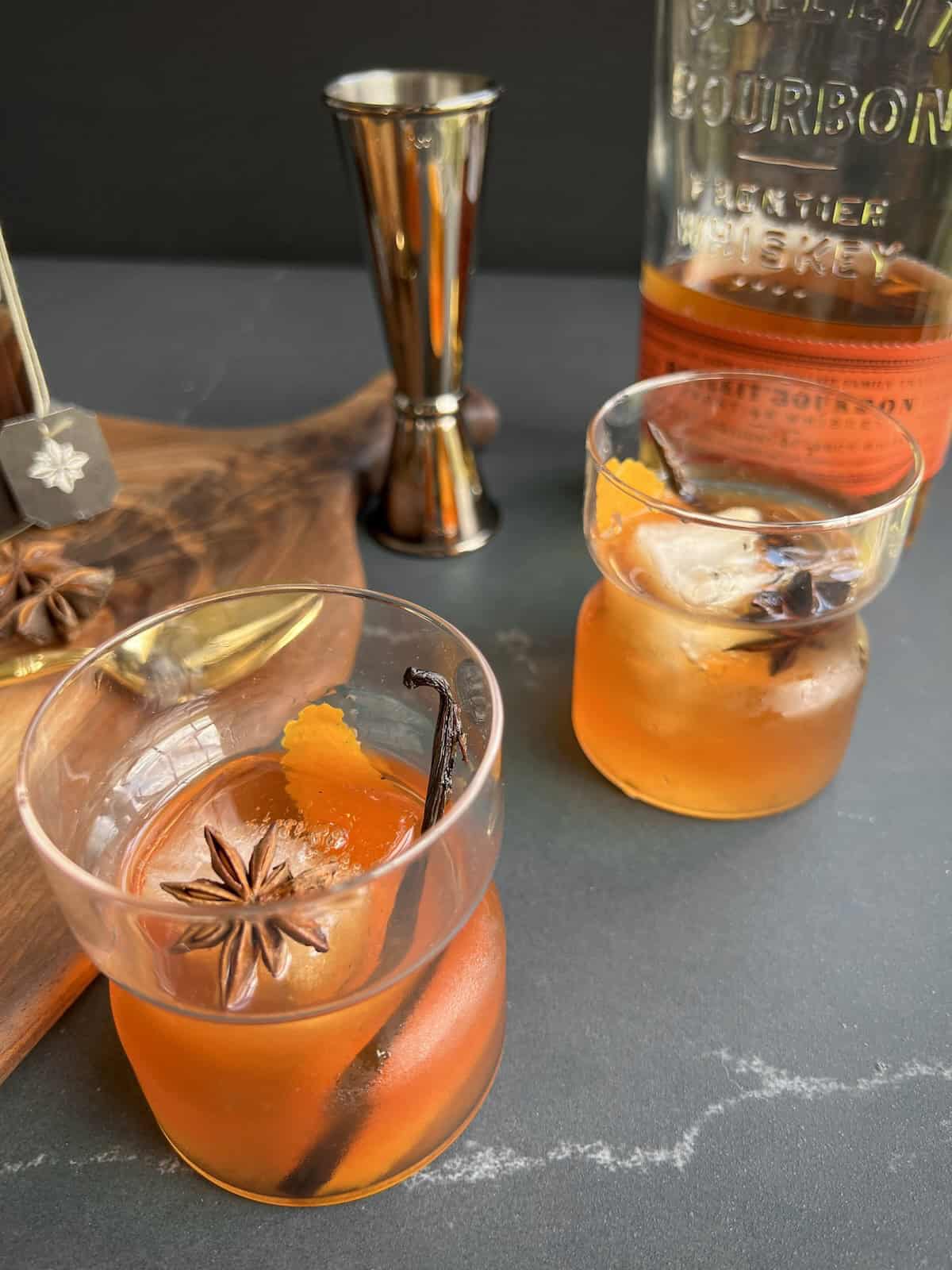 two vanilla chai old fashioned cocktails sit on countertop with a jigger and bottle of whiskey in the background