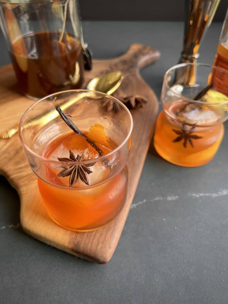 a rocks glass containing a finished vanilla chai old fashioned is served on wooden board surrounded by bar tools and bottles.