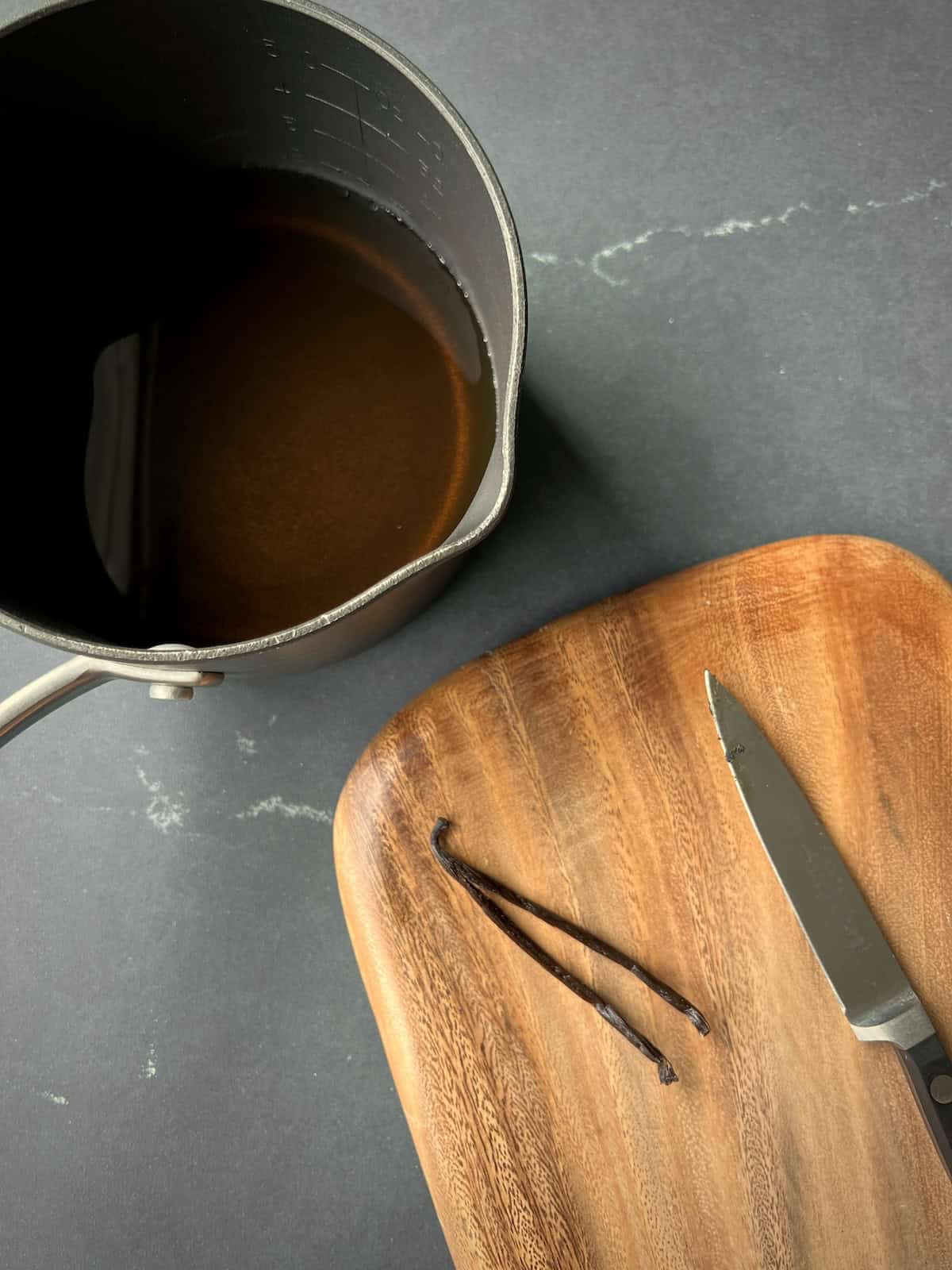 a halved vanilla bean is sliced lengthwise on a wooden cutting board next to a small saucepan of liquid.