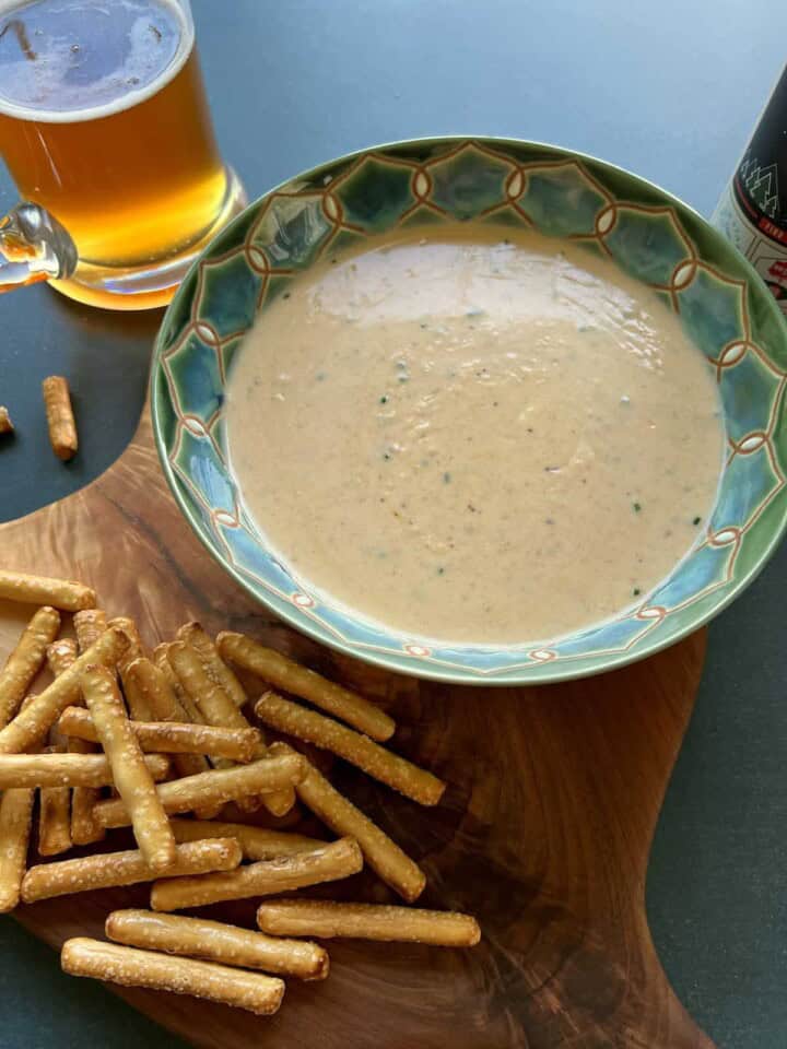a bowl of oktoberfest beer cheese sauce sits on a wooden board next to a pile of pretzels and a mug of golden hued beer