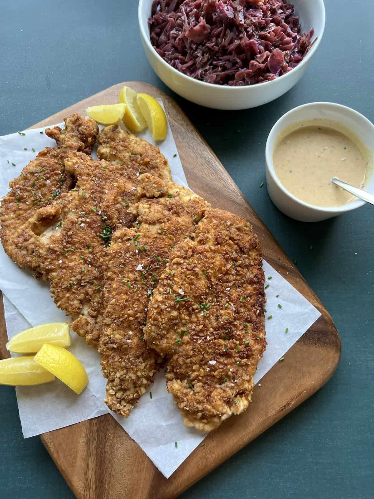 the cooked pretzel crusted chicken schnitzel pieces are arranged on parchment paper on a wooden board and garnished with minced chives, flaky salt, and lemon wedges. 