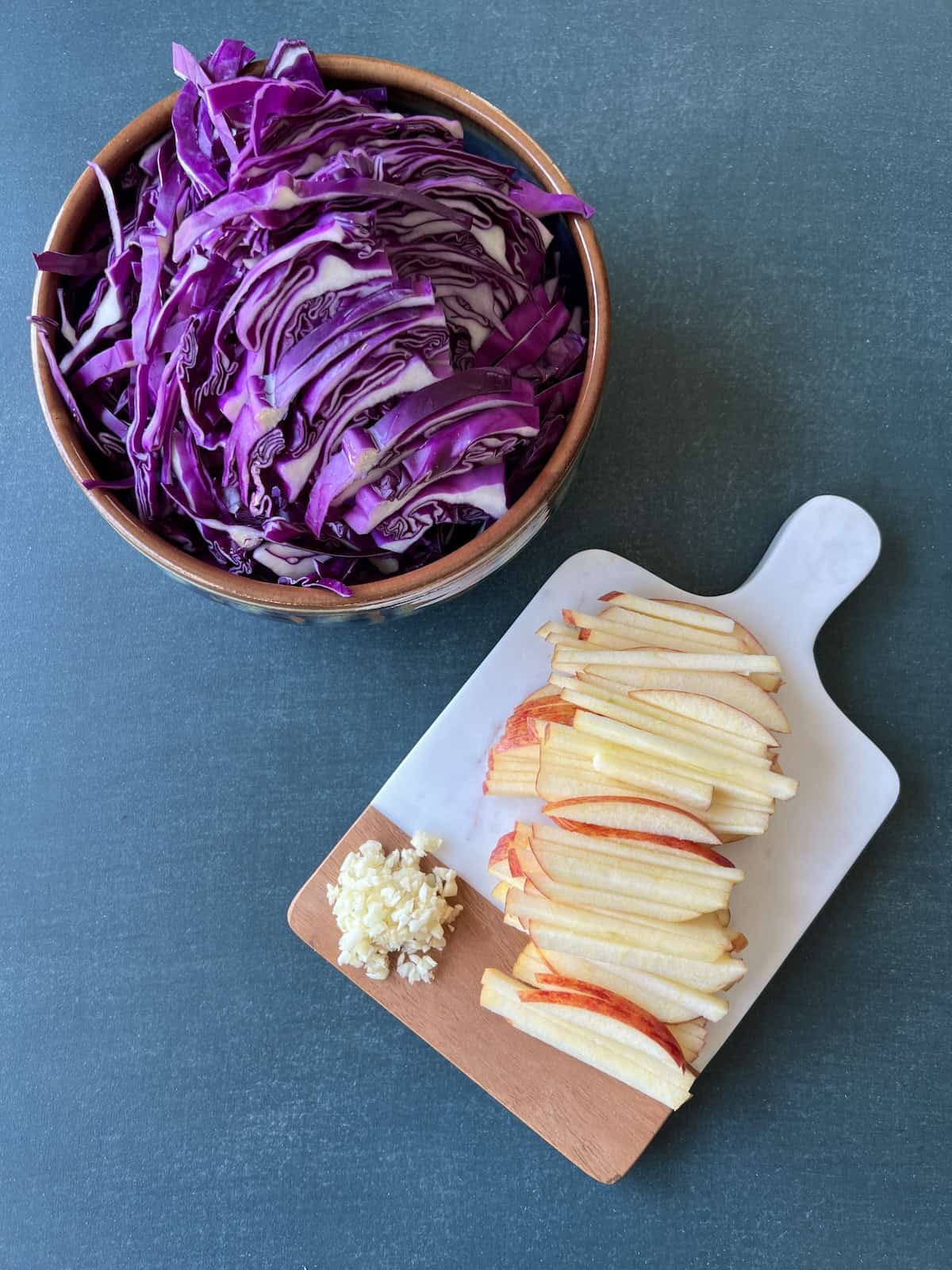 a bowl of sliced red cabbage is sitting next to a small cutting board with sliced apple and minced garlic on it.