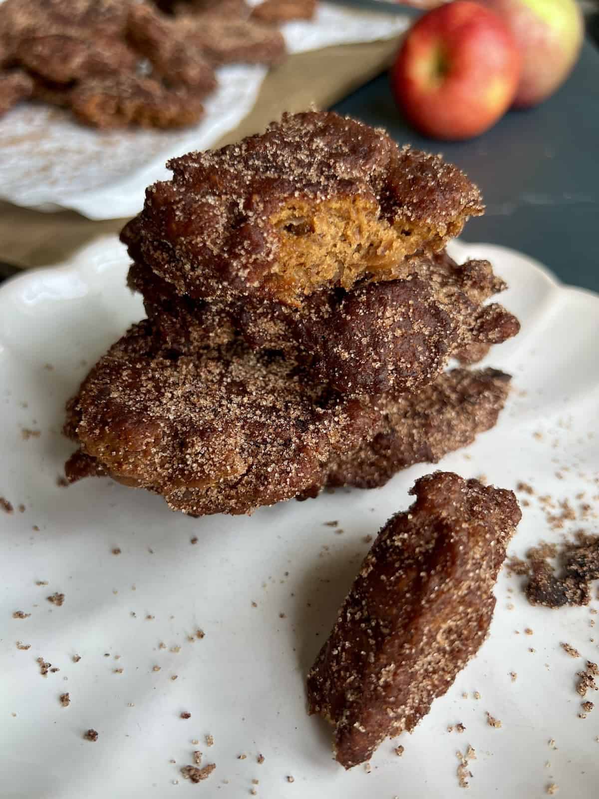 a stack of 3 applesauce fritters sits in front of a wooden board with more cooked fritters and a couple of fresh red apples