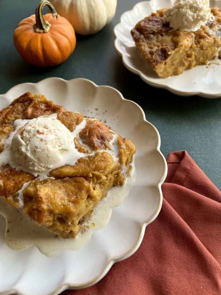 two plates of pumpkin brioche bread pudding are topped with scoops of vanilla ice cream to serve