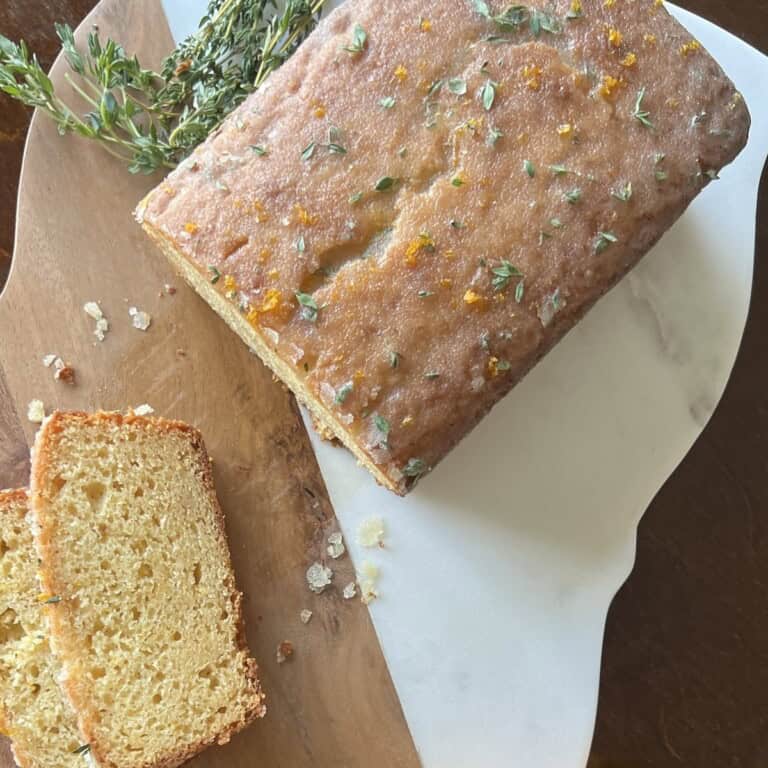 whole meyer lemon cake sits on a cutting board. 2 slices have been cut off and fresh thyme sprigs and whole lemons are nearby.