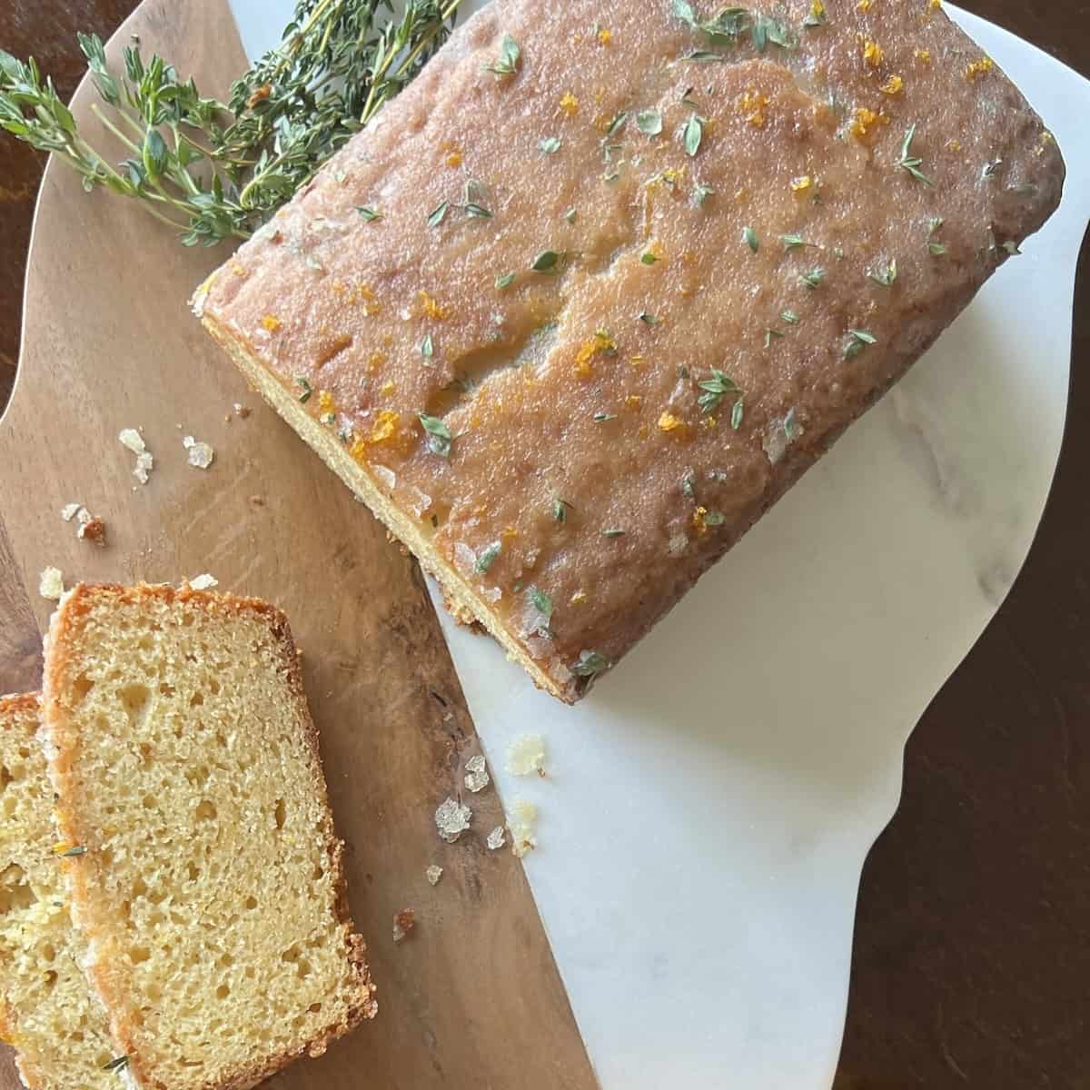 whole meyer lemon cake sits on a cutting board. 2 slices have been cut off and fresh thyme sprigs and whole lemons are nearby.