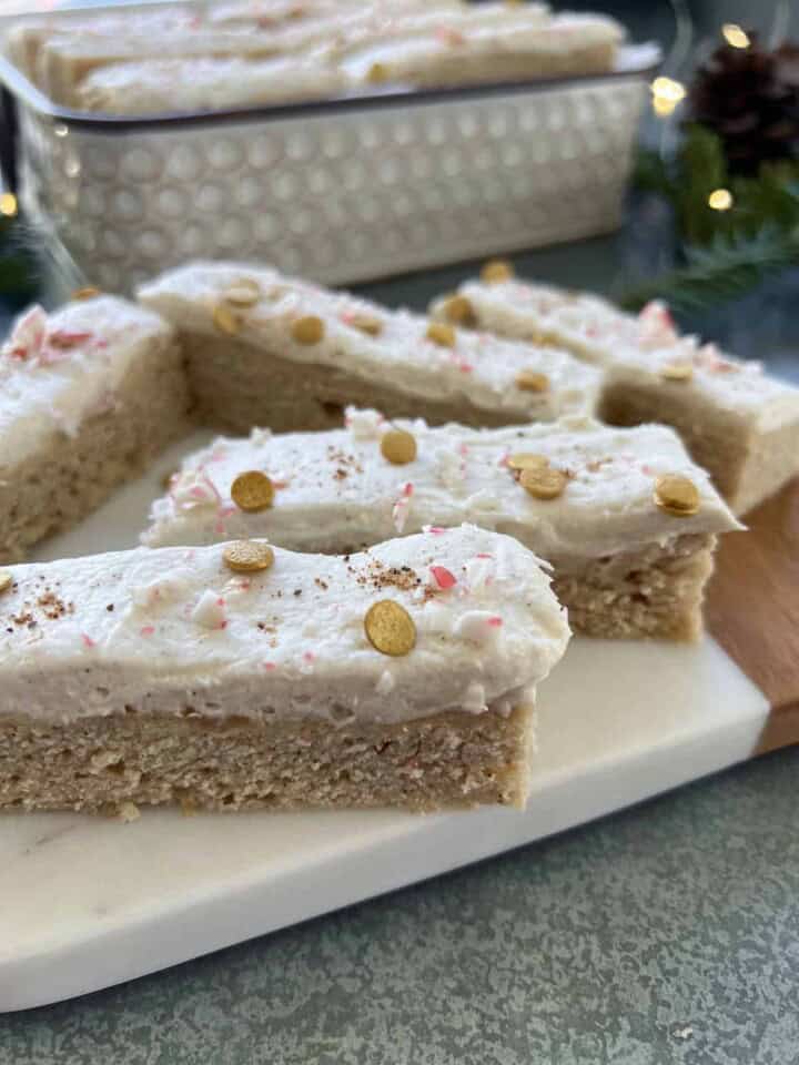 eggnog cookie bars on a small serving board sit in front of a white tin filled with more cookie bars