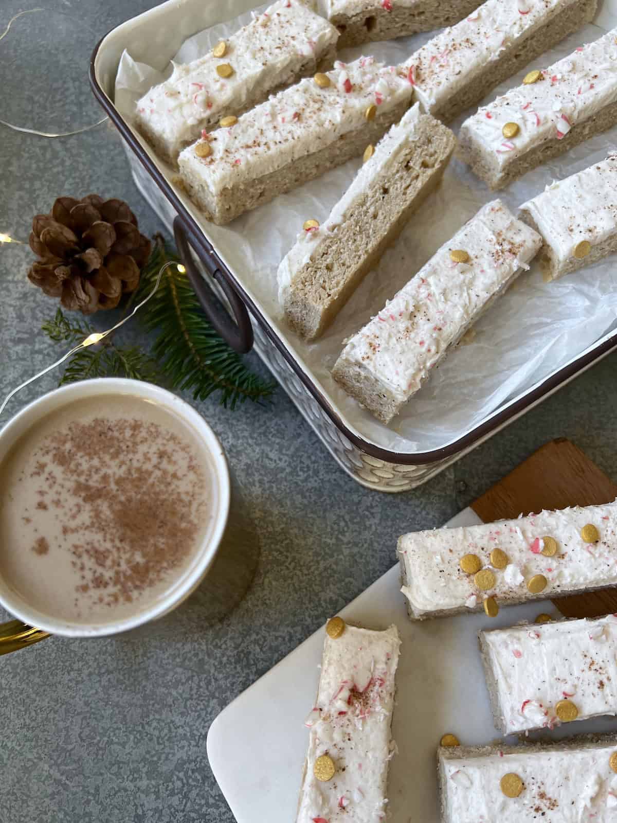 eggnog cookie bars are lined up in a white tin and on a small platter, set on a table next to a gold mug of coffee