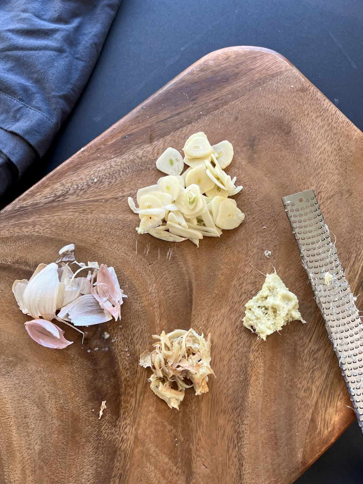 a pile of grated fresh ginger and sliced garlic cloves sit on a wooden cutting board, with their respective peels discarded nearby