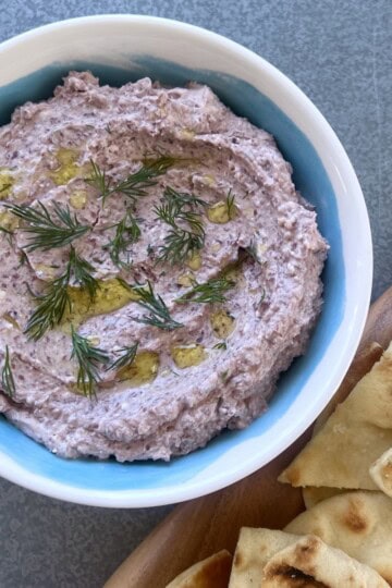 a bowl of mediterranean olive dip is garnished with fresh dill and set next to a serving board of sliced pita.