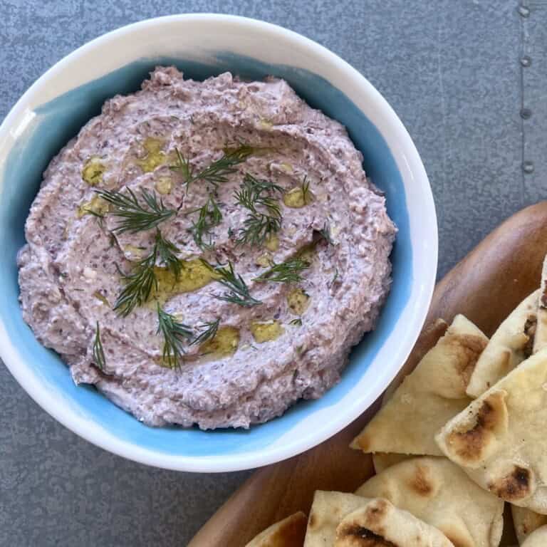 a bowl of mediterranean olive dip is garnished with fresh dill and set next to a serving board of sliced pita.