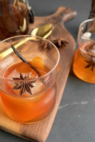 a rocks glass containing a finished vanilla chai old fashioned is served on wooden board surrounded by bar tools and bottles.