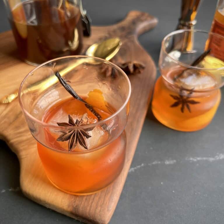 a rocks glass containing a finished vanilla chai old fashioned is served on wooden board surrounded by bar tools and bottles.