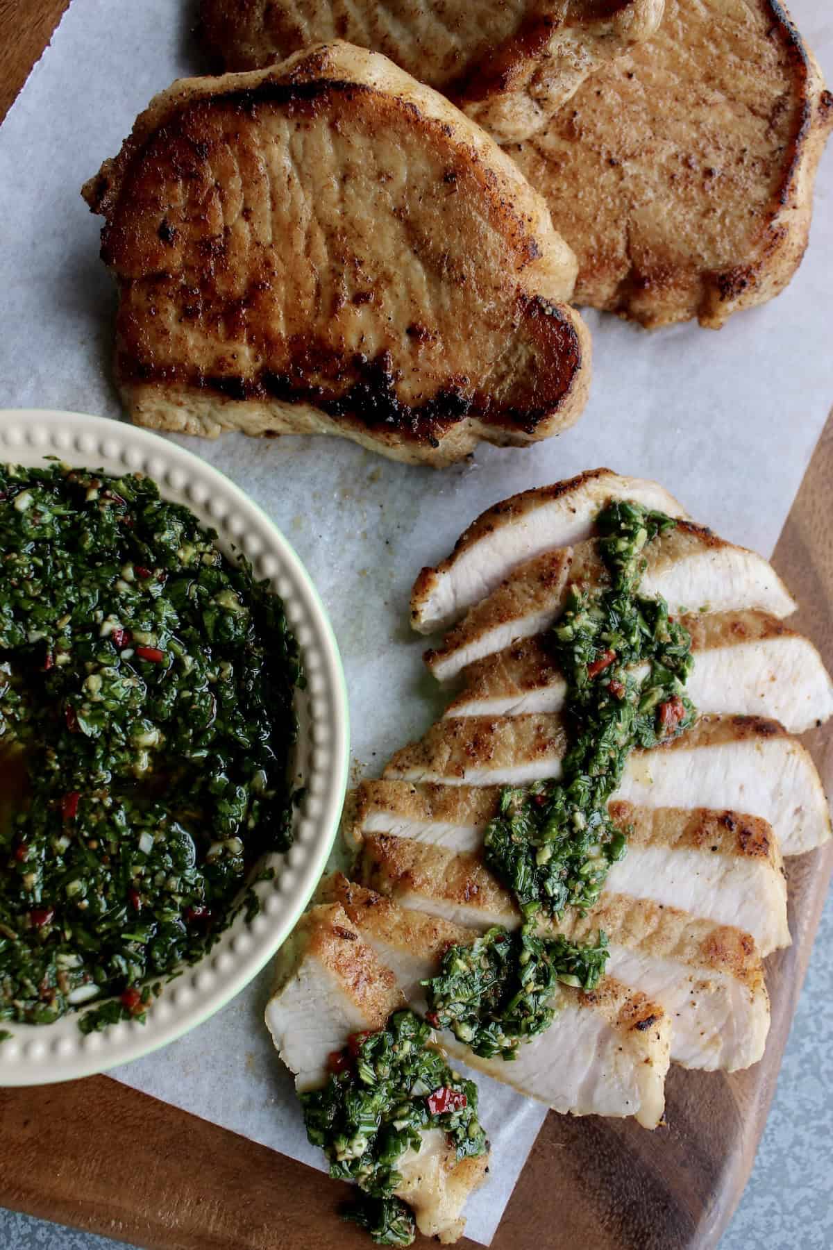 a pork chop is sliced on a  wooden board is topped with a line of chimichurri while the 3 whole pork chops rest nearby