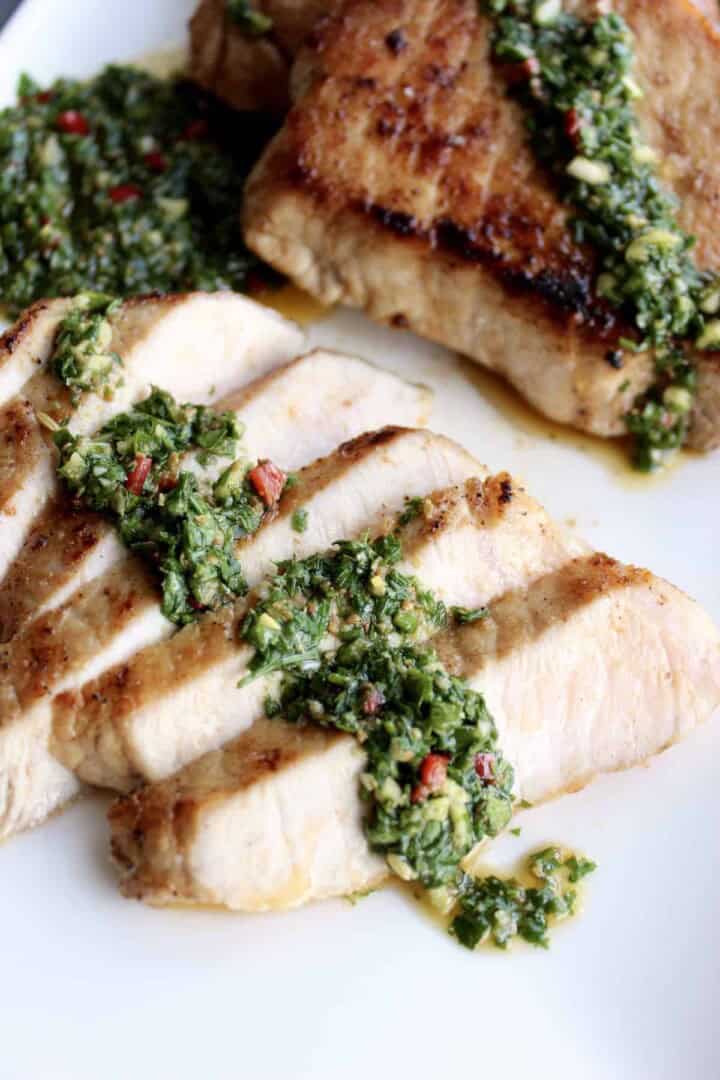 slices of a chimichurri pork chop are fanned out on a plate topped with a line of bright green chimichurri sauce