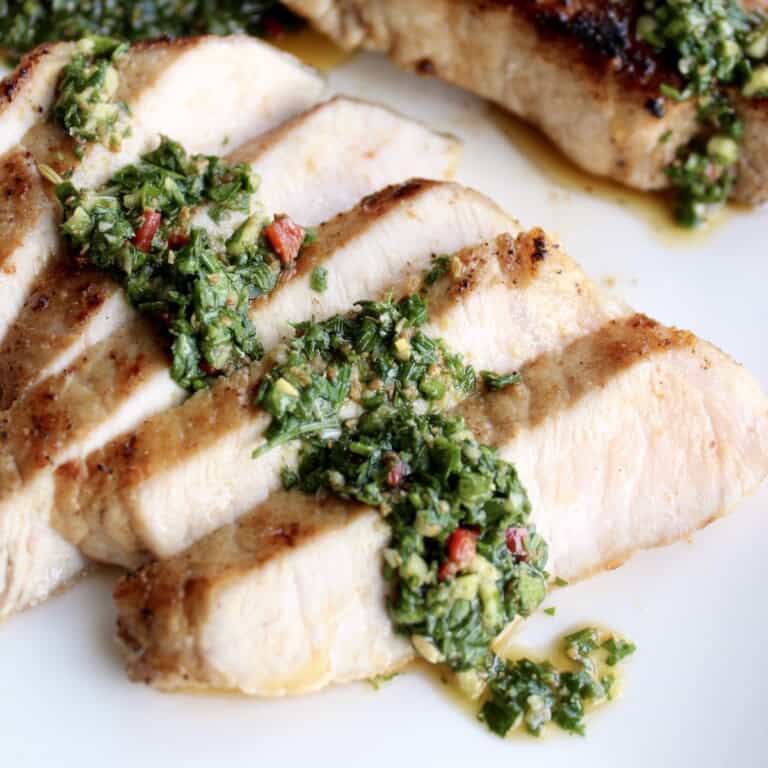 slices of a chimichurri pork chop are fanned out on a plate topped with a line of bright green chimichurri sauce
