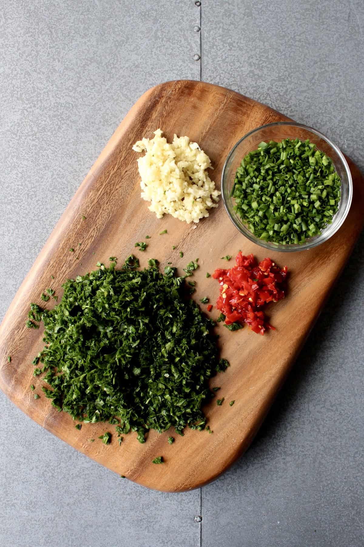 small piles of minced parsley, chives, garlic, and red chili pepper are arranged on a wooden cutting board