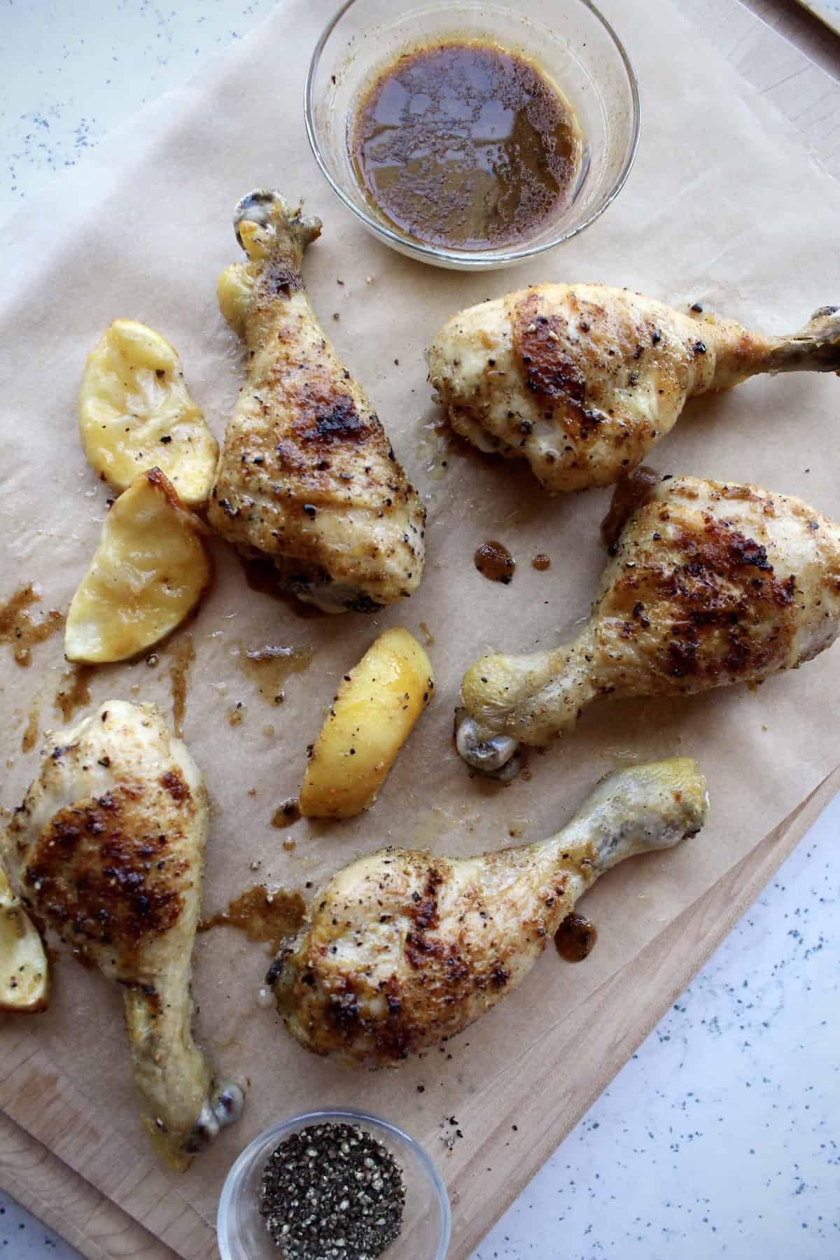 lemon pepper chicken legs are served on a wooden board with a small bowl of pan drippings and addition cracked black pepper