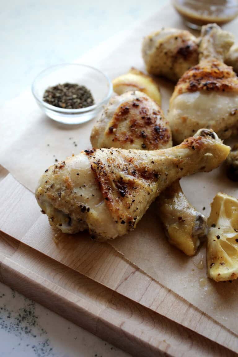 crispy lemon pepper chicken legs are piled on a wooden board and served with caramelized lemon wedges and cracked black pepper