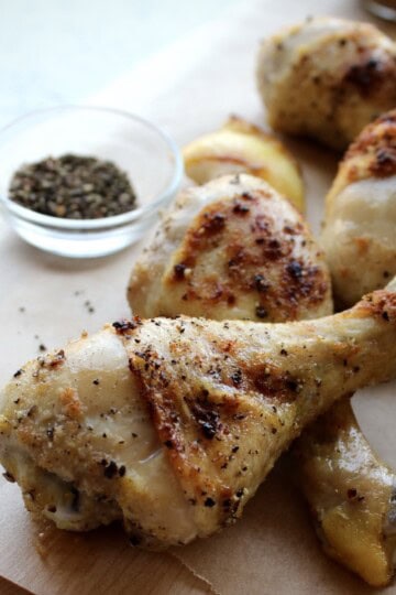crispy lemon pepper chicken legs are piled on a wooden board and served with caramelized lemon wedges and cracked black pepper