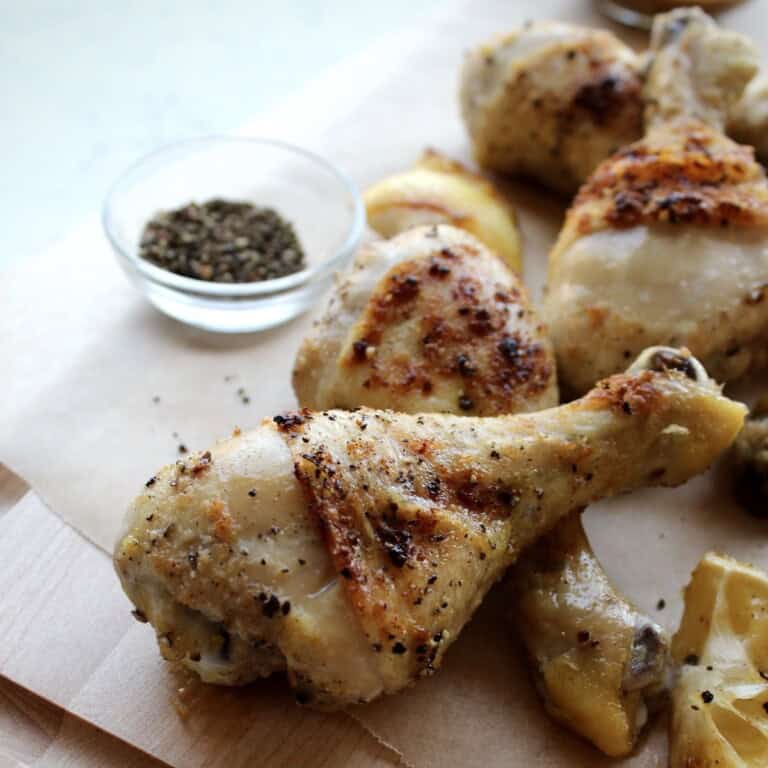 crispy lemon pepper chicken legs are piled on a wooden board and served with caramelized lemon wedges and cracked black pepper