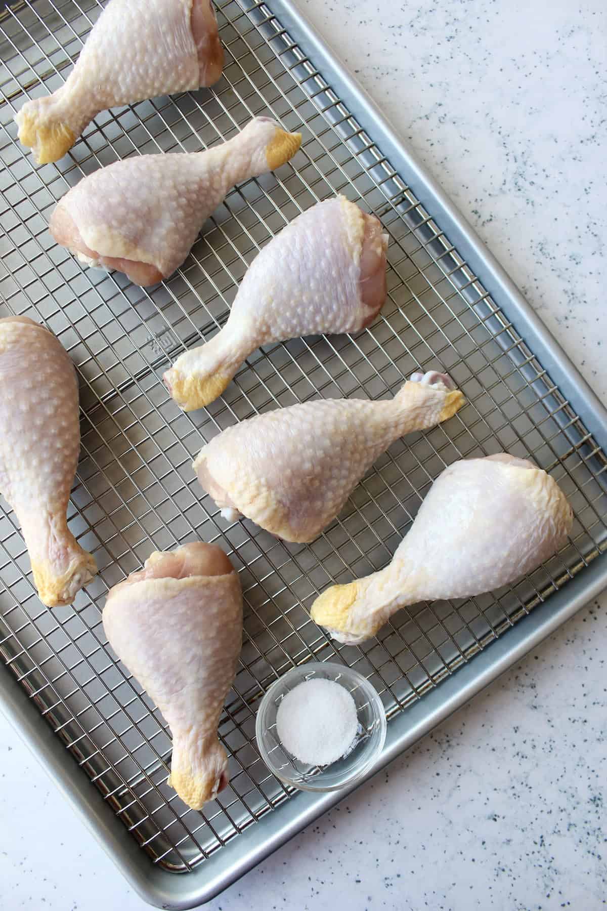 raw chicken legs are arranged on a baking sheet with a wire rack next to a small bowl of salt