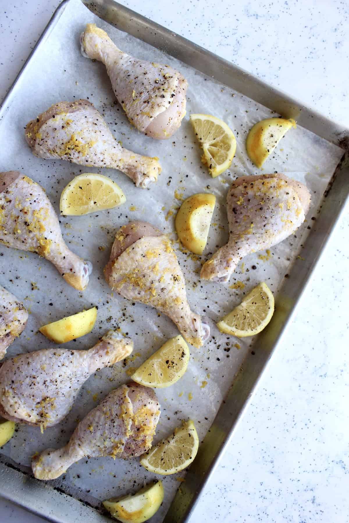 seasoned chicken legs and thick lemon wedges are arranged on a parchment paper lined baking sheet.